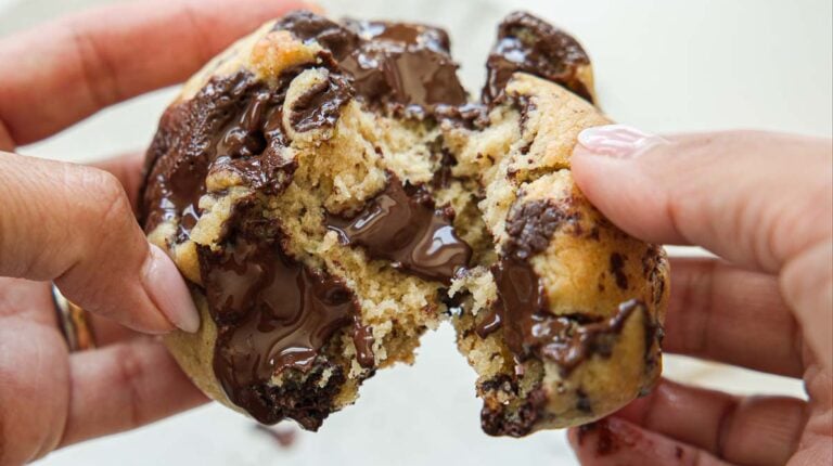 Close-up of hands pulling apart a warm, gooey vegan chocolate chip cookie, with melted chocolate oozing from the center.